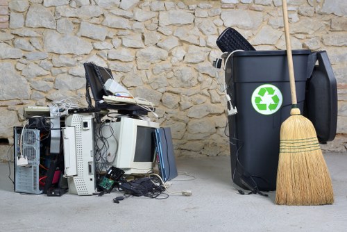 A collection of discarded electronic waste, including computer monitors, keyboards, and various small hardware components, is stacked on the ground next to a large black rubbish bin with a green recycling symbol. The electronic items display different textures such as glossy screens, matte plastic casings, and metallic parts, with some cables and wires visible among the debris. The rubbish bin is positioned against a stone wall, and a wooden broom with stiff bristles leans against it. The scene is outdoors, on a concrete surface, suggestive of a waste collection or disposal area in an urban setting. The presence of the rubbish bin indicates a waste management context, typical of a commercial or residential waste removal service operating in Poplar, possibly handled by Commercial Waste Poplar. The environment is well-lit, with natural daylight illuminating the scene, emphasizing the contrast between the electronic waste and the surrounding environment, supporting the context of rubbish removal services for electronics and general waste disposal.