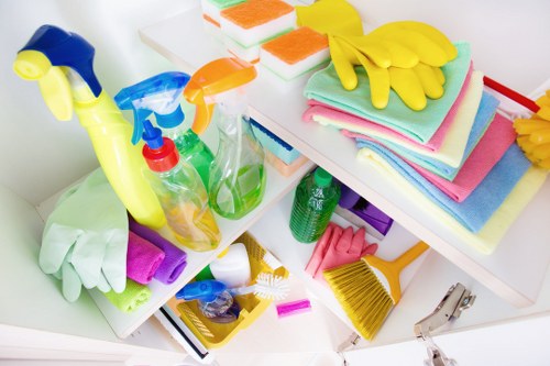 The image displays a cluttered countertop or shelf with various cleaning and hygiene supplies, including multiple spray bottles in green, orange, and blue with spray nozzles, stacks of folded cleaning cloths in pastel shades such as pink, blue, and white, and a pair of bright yellow rubber gloves resting on top of the cloths. Adjacent to the cloths are cleaning tools like a small yellow-handled brush and a yellow dustpan with a brush, as well as some disposable sponges with orange and green surfaces. The background features additional cleaning accessories, and the overall scene suggests an organized area for domestic or commercial cleaning tasks, typical of waste disposal preparation in a Poplar-based cleaning or rubbish removal context, as managed by companies like Commercial Waste Poplar. The setting's bright lighting highlights the vibrant colors and clean textures of the various cleaning products and tools, emphasizing their readiness for use in waste management or cleaning projects within the local area.