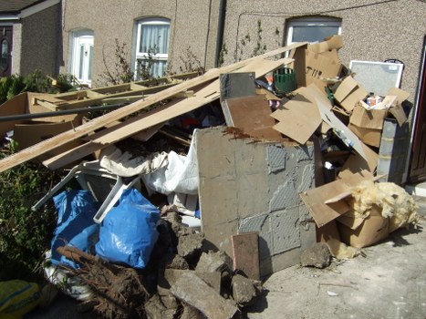 An outdoor pile of mixed waste and debris located on a paved driveway in a residential area, with a backdrop of a beige residential building featuring white-framed windows. The rubbish includes large cardboard boxes, some flattened and some intact, with visible corrugated edges and brown kraft paper surfaces, as well as broken wooden planks with a pale, unfinished appearance leaning against the heap. There are also pieces of concrete and stone rubble with rough, uneven textures and grey coloration, and a white plastic bag partially visible beneath the debris. The waste appears haphazardly arranged, casting shadows on the ground, suggesting natural sunlight. The scene shows an accumulation of discarded household and building materials, indicative of a waste removal service organized by companies like Commercial Waste Poplar in the East London area, near Poplar postal district, supporting local waste management needs.
