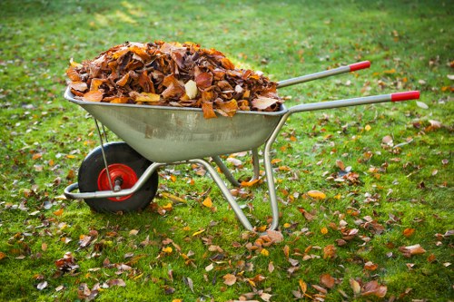 A silver metal wheelbarrow with red handles and a black wheel, filled with a large quantity of brown and yellow autumn leaves, situated on a grassy lawn scattered with fallen leaves. The wheelbarrow is positioned in the foreground with a green, slightly blurred background indicating an outdoor setting, possibly a garden or yard. The scene depicts a typical leaf clearing activity that relates to rubbish removal services offered by Commercial Waste Poplar in the Poplar area of London, with the leaves representing garden or seasonal waste collected for disposal or collection.