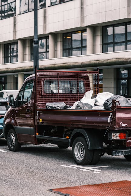 A small burgundy pickup truck parked on an urban street in front of a modern office building with large glass windows. The truck's open flatbed is filled with various types of collected rubbish, inclu
