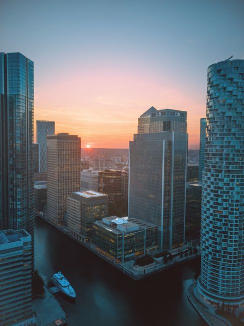 A panoramic view of a modern urban waterfront with several high-rise office buildings and skyscrapers. The buildings feature glass facades with reflective surfaces, illuminated windows, and metallic f